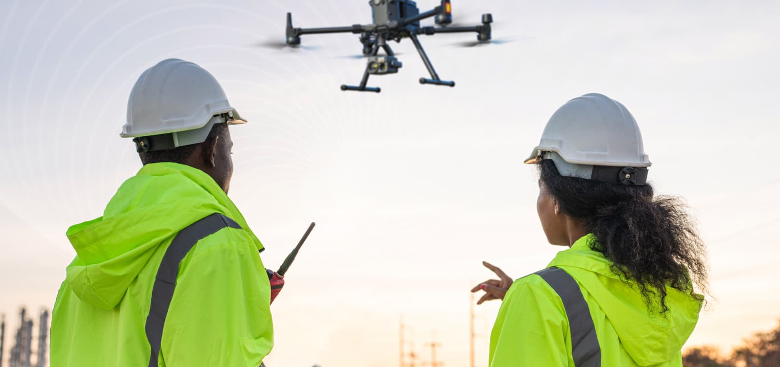 Two people in high vis jackets observing an enterprise-grade DJI M30 drone, with the person on the right pointing at the drone, with a background of a remote site facility. 